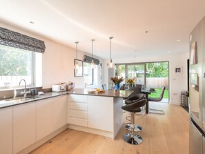 A modern kitchen with a central island, bar stools, a dining area, and a view of the backyard. - Pentire (Wadebridge)