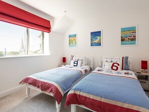A bedroom with two beds, a window with red curtains, and three framed pictures on the wall. - Pentire (Wadebridge)