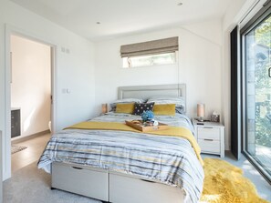 A modern bedroom with a large bed, bedside tables, a window with a view, and a door leading to another room. - Pentire (Wadebridge)