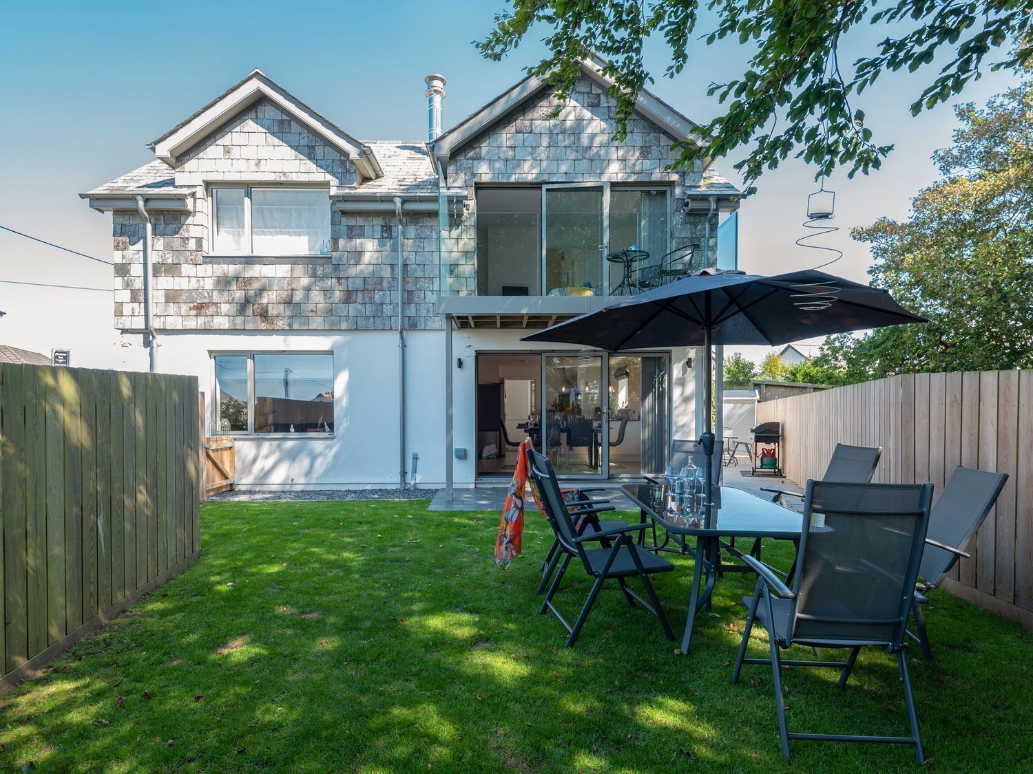 Een modern huis met twee verdiepingen en een patio met een tafel, stoelen, een grote parasol en een houten omheining.