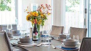 Una mesa de comedor dispuesta con platos, tazones, cubiertos y copas de vino, con un jarrón de flores en el centro.