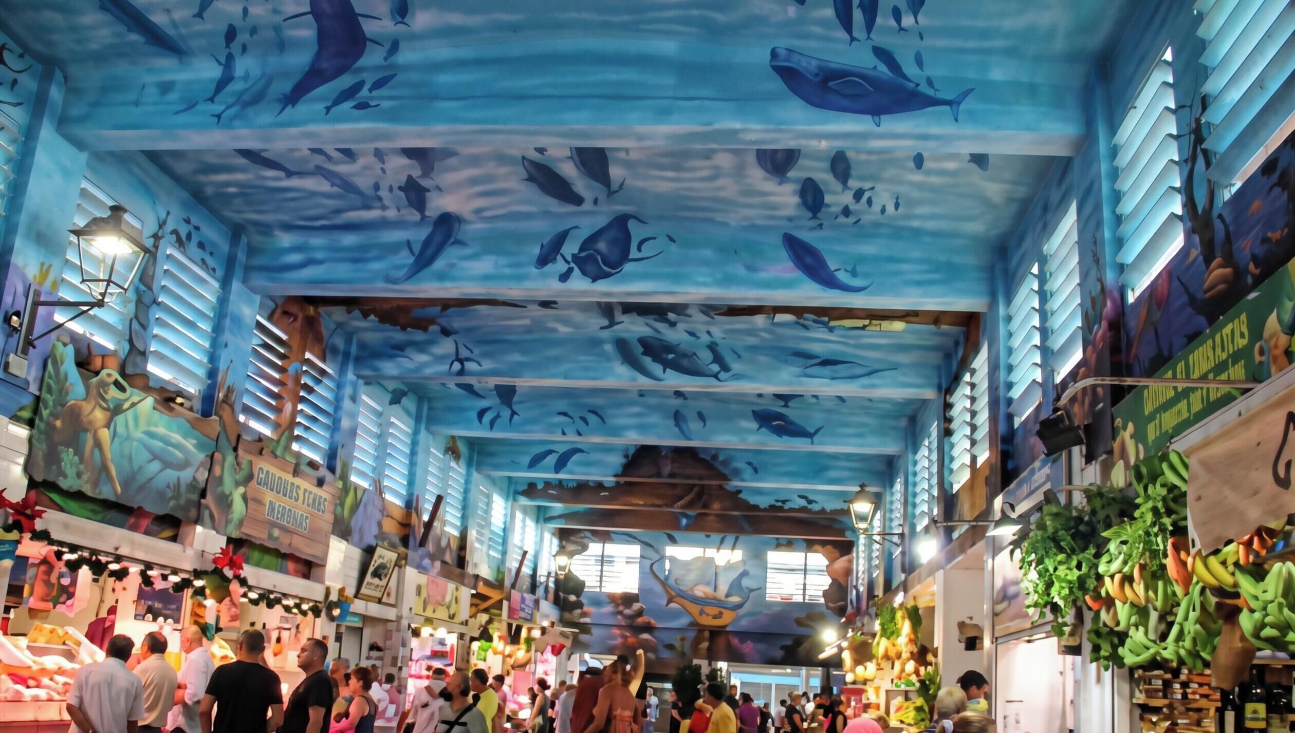 An indoor market with a ceiling mural of marine life, wooden beams, and various stalls selling fruits and vegetables.