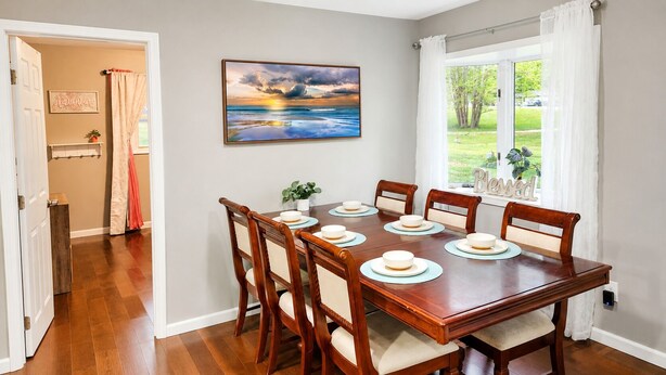 Comedor con una mesa de madera dispuesta para seis comensales, un cuadro de un atardecer y vistas a un jardín desde una ventana.