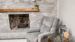 Una habitación con chimenea de ladrillo, un sillón gris y una mesa de vidrio con libros y objetos decorativos.