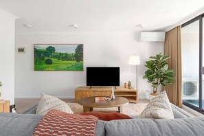 Ein modernes Wohnzimmer mit einem Flachbildfernseher, einem runden Couchtisch, einer Couch und einem großen Fenster mit Vorhängen.