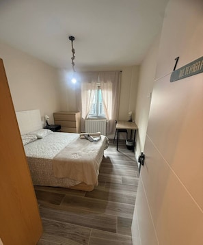 A bedroom with a bed, a desk, a chair, a radiator, and a window with curtains.