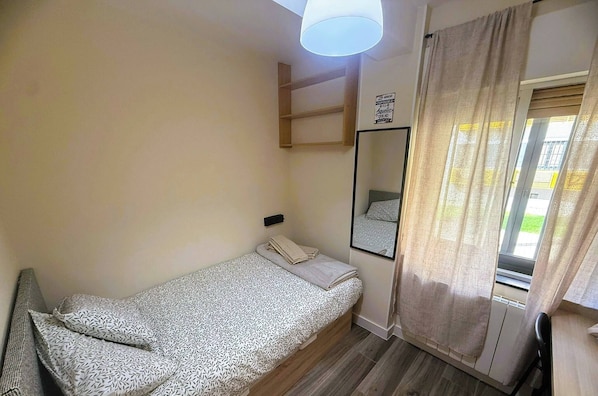 A single bed with a patterned bedspread, a bedside table, a mirror, a shelf, and a window with curtains.