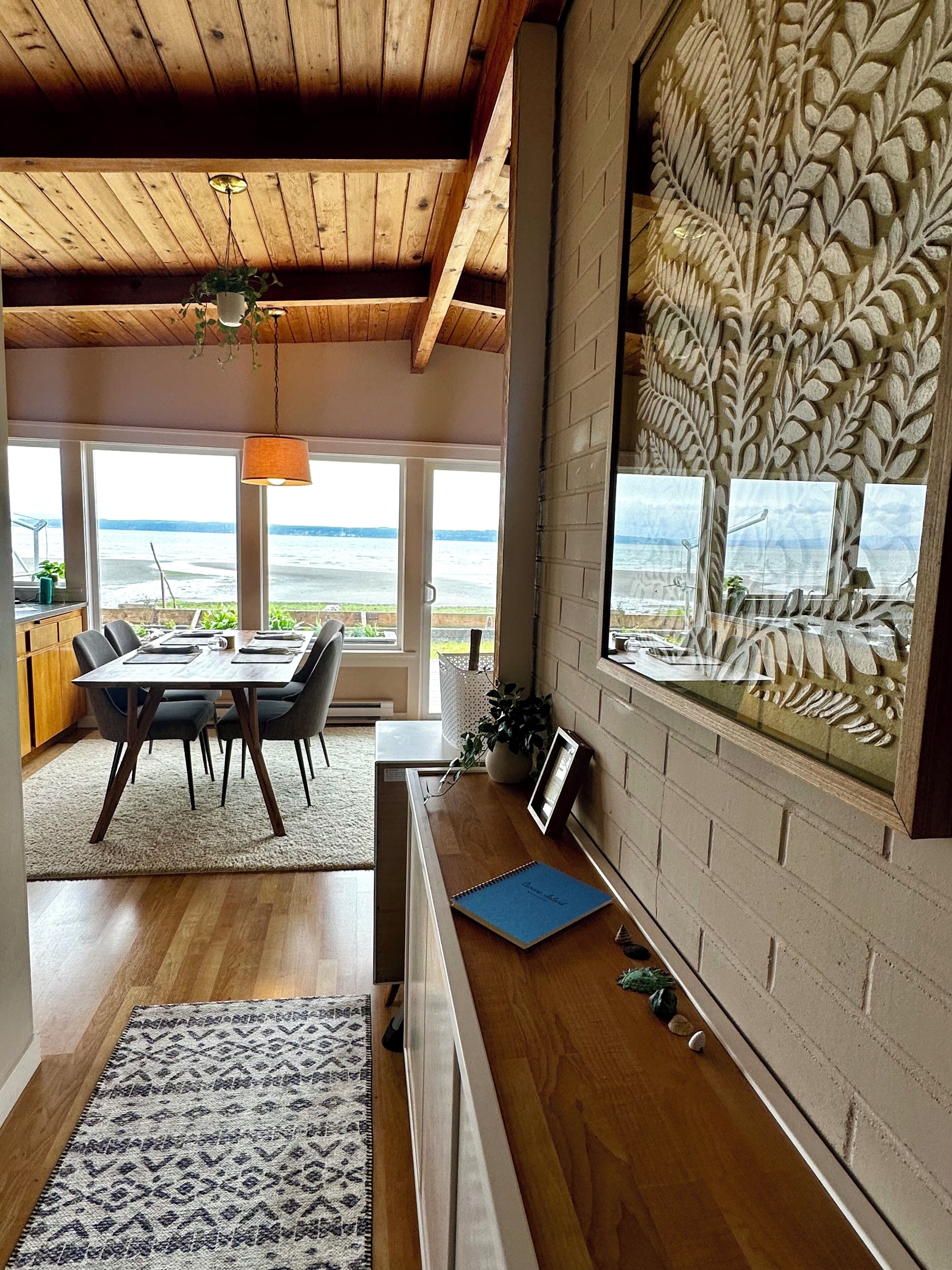 Un intérieur moderne avec un plafond en bois, un coin repas offrant une vue sur la mer et un motif décoratif en forme de feuilles sur le mur.