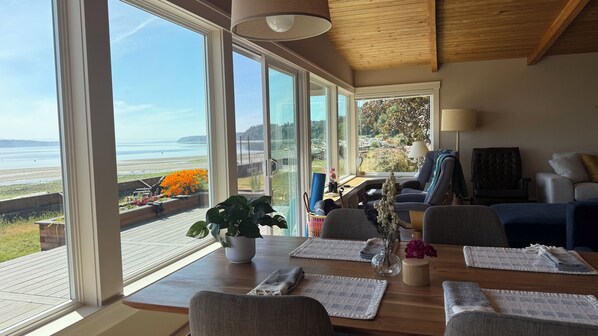 A modern living room with large windows, a wooden dining table, and a view of the ocean. - Enjoy Camano Tidalwilds waterfront beach house w/ hot tub, sups, kayak and more! (Camano)
