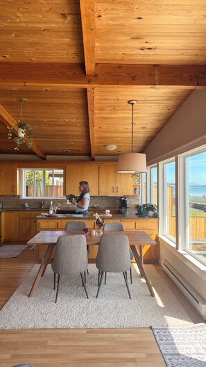 A modern kitchen with wooden cabinetry, a large window, and a dining area with a table and chairs. - Enjoy Camano Tidalwilds waterfront beach house w/ hot tub, sups, kayak and more! (Camano)