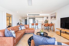 Ein modernes Wohnzimmer mit einer Couch, einem Couchtisch und einem Fernseher. Im Hintergrund befindet sich ein Essbereich mit Stühlen und eine Küche.