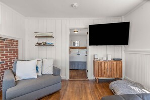 A room with a grey sofa, a wooden cabinet, a flat-screen TV, and a bed in the background.
