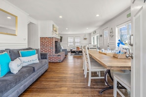 A modern living room with a fireplace, a dining area with a wooden table and chairs, and a grey sofa with blue cushions.