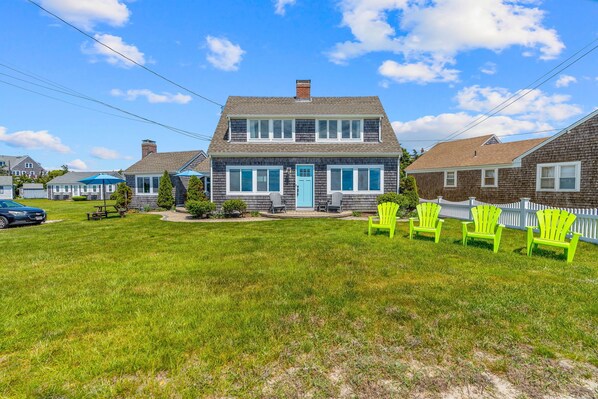 A two-story house with a front porch, a blue door, and a lawn with green chairs.