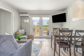 Un salon comprenant un canapĂ©, une table Ă manger avec des chaises, un tĂ©lĂ©viseur fixĂ© au mur et une porte-fenĂȘtre coulissante donnant sur un espace extĂ©rieur.