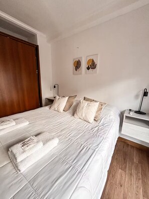 Un lit bien fait, avec du linge de lit et des serviettes blanches, une porte en bois, une table de chevet avec une lampe et deux œuvres d’art accrochées au mur.