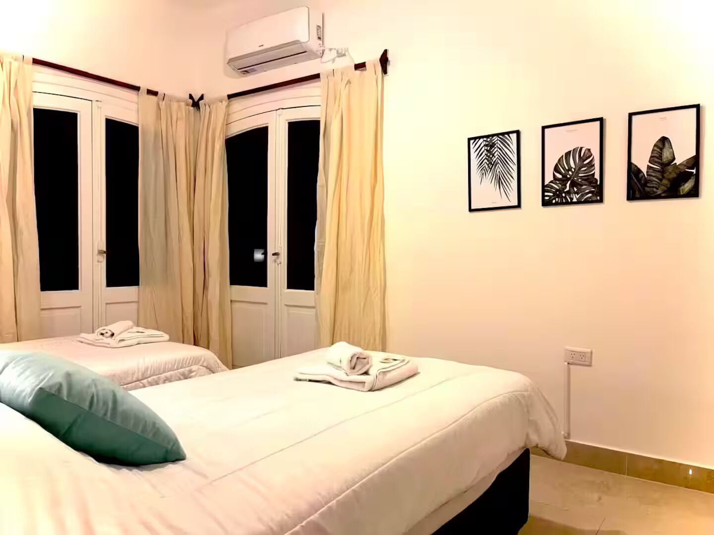 Camera d'albergo con due letti, una finestra con tende, un condizionatore d'aria e tre opere d'arte con foglie in cornice appese al muro.