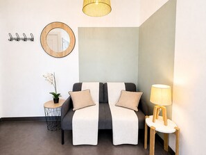 Een moderne kamer met een grijze bank, een klein rond tafeltje met een lamp, een spiegel en een kapstok aan de muur.