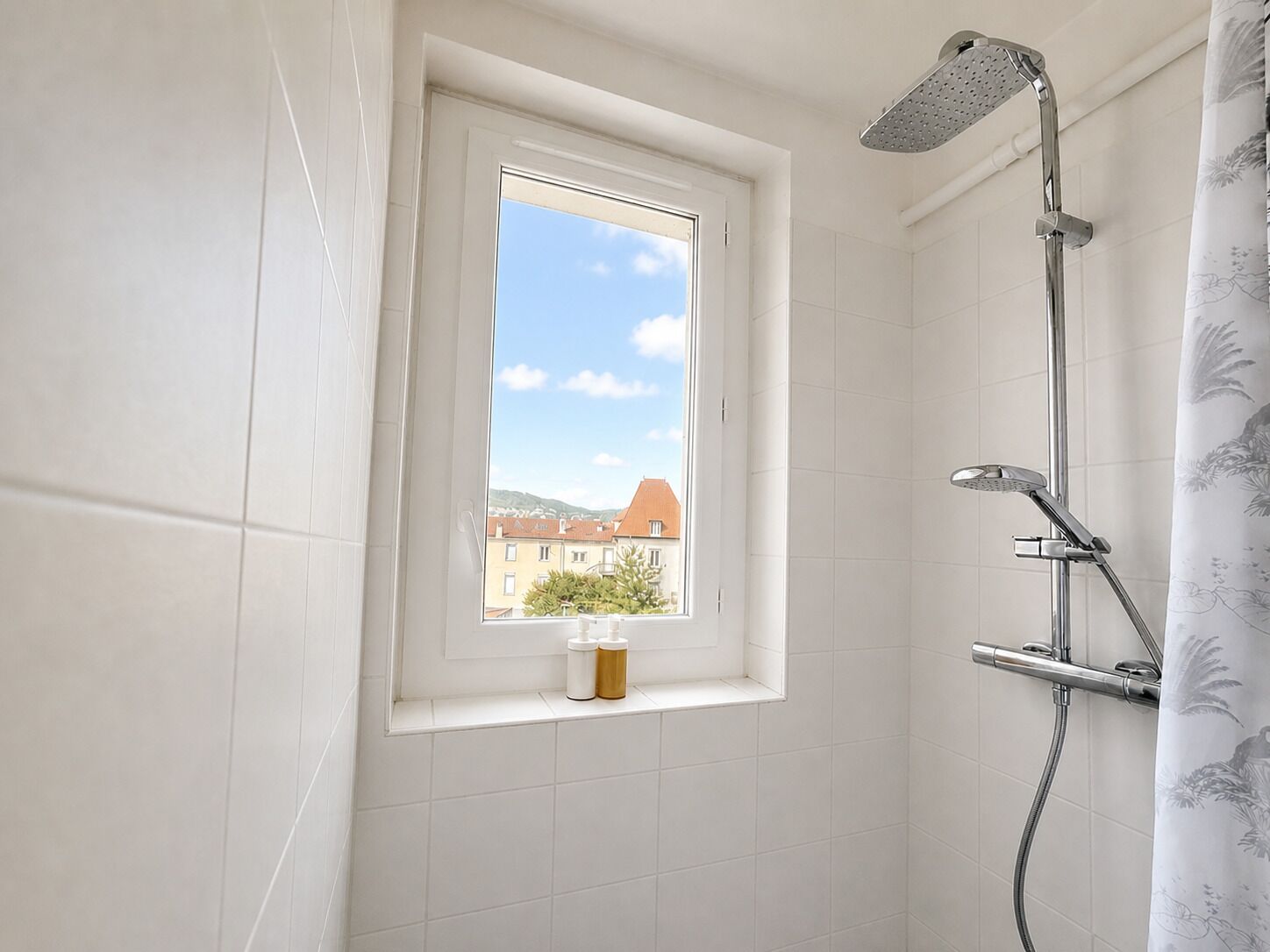 Ein Badezimmer mit Dusche, einem Fenster mit Blick auf Gebäude und einem Regal mit Flaschen.