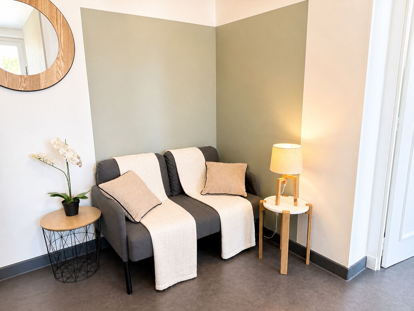 A corner room with a grey sofa, two pillows, a small round table with a lamp, a potted plant, and a round mirror on the wall.