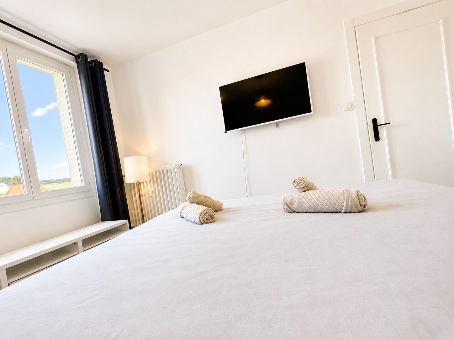 A hotel room with a large bed, a TV mounted on the wall, a window with curtains, and a radiator.