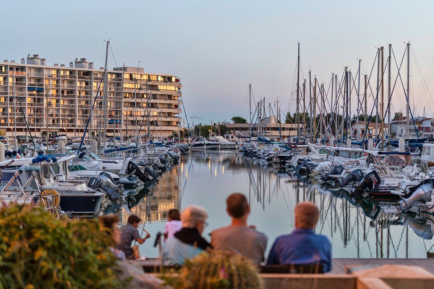 A marina with boats docked, a building with balconies, and people seated on a deck.