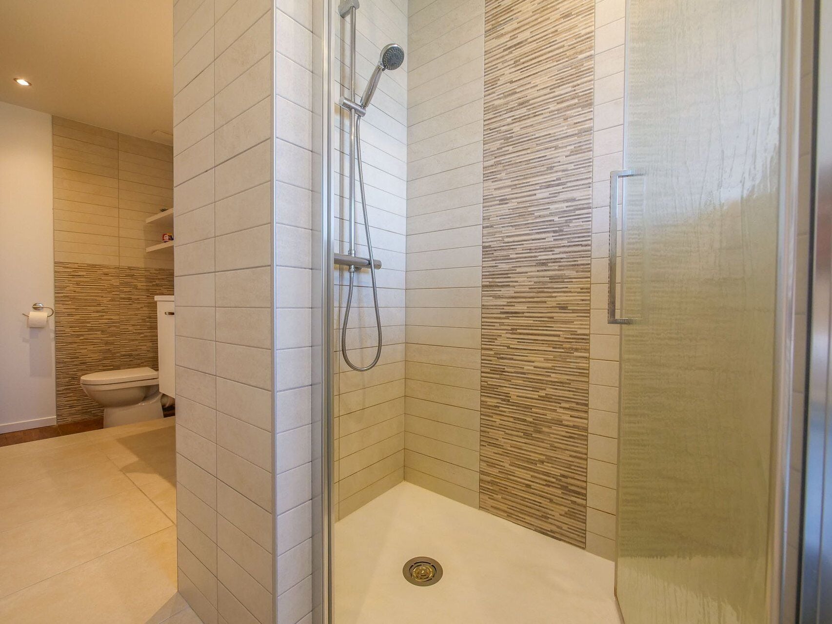 Een moderne badkamer met een glazen douchecabine, betegelde wanden en een toilet.