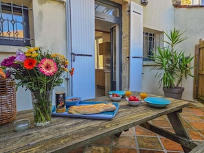 Una mesa de madera con alimentos para el desayuno, un jarrón de flores y una planta en maceta en un patio.