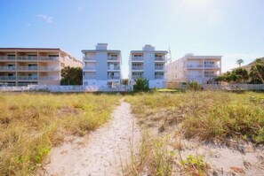 Ein Weg führt zu einem Hotel direkt am Strand, mit mehreren Balkonen und einem Sandstrand im Vordergrund.