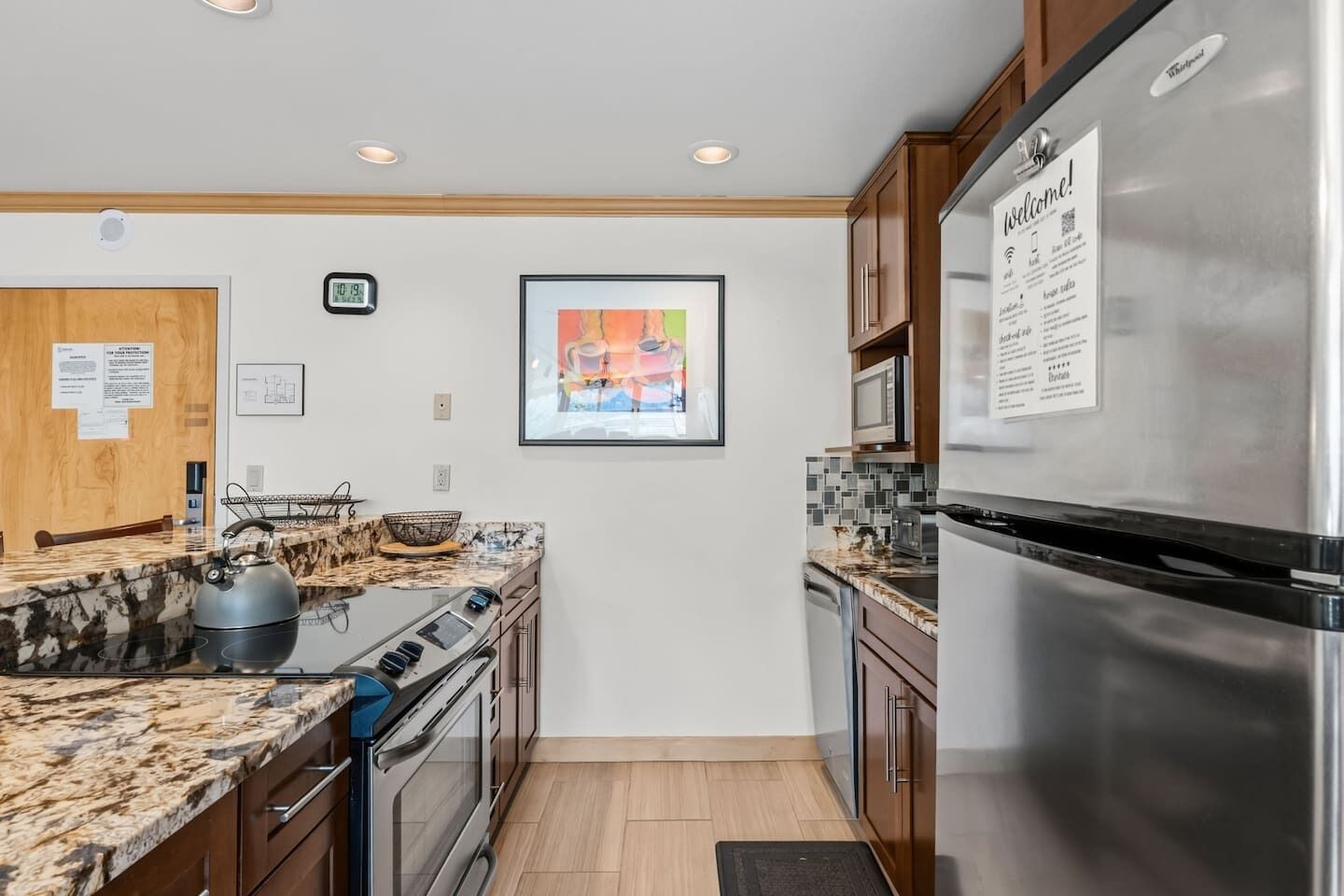 A modern kitchen with stainless steel appliances, a marble countertop, and a framed artwork on the wall.