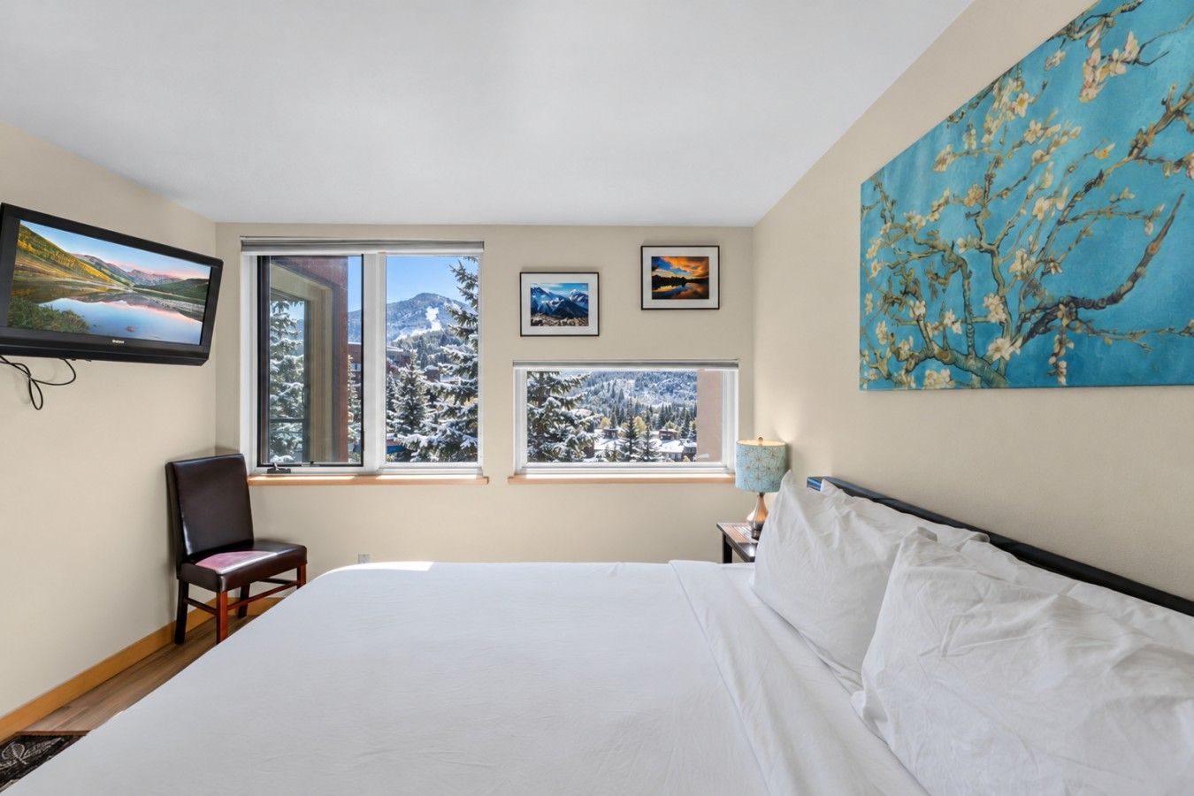 A hotel room with a large bed, two windows with snowy views, a chair, and artwork on the walls.