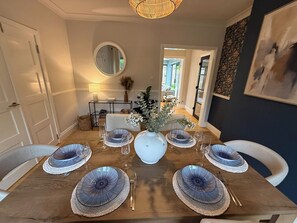 Ein Esszimmer mit einem Holztisch, gedeckt für sechs Personen, weißen Stühlen, einer Vase mit Blumen und einem Spiegel an der Wand.