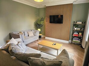 A living room with a sofa, a coffee table, a TV mounted on a wooden wall, and a bookshelf with board games. - Save 25% OFF Monthly| Sharrow Retreat | Sleeps 4 (North Yorkshire)