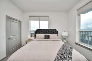 Habitación de hotel con una cama grande, ventana con vista a la ciudad, puerta y mesita de noche con lámpara.