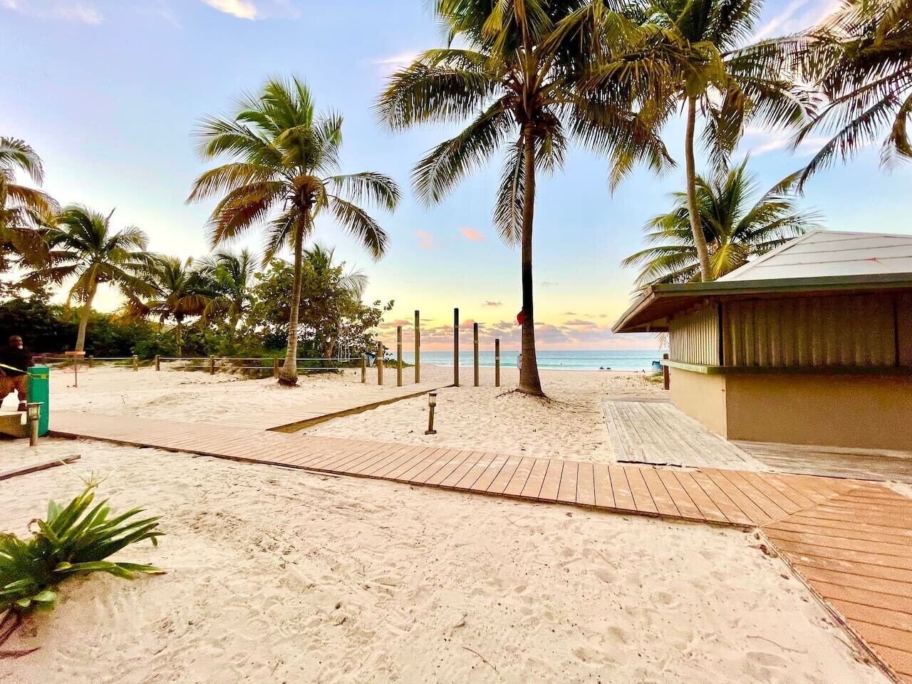 Ein Strand mit Palmen, einem hölzernen Steg und einem Gebäude mit geneigtem Dach.