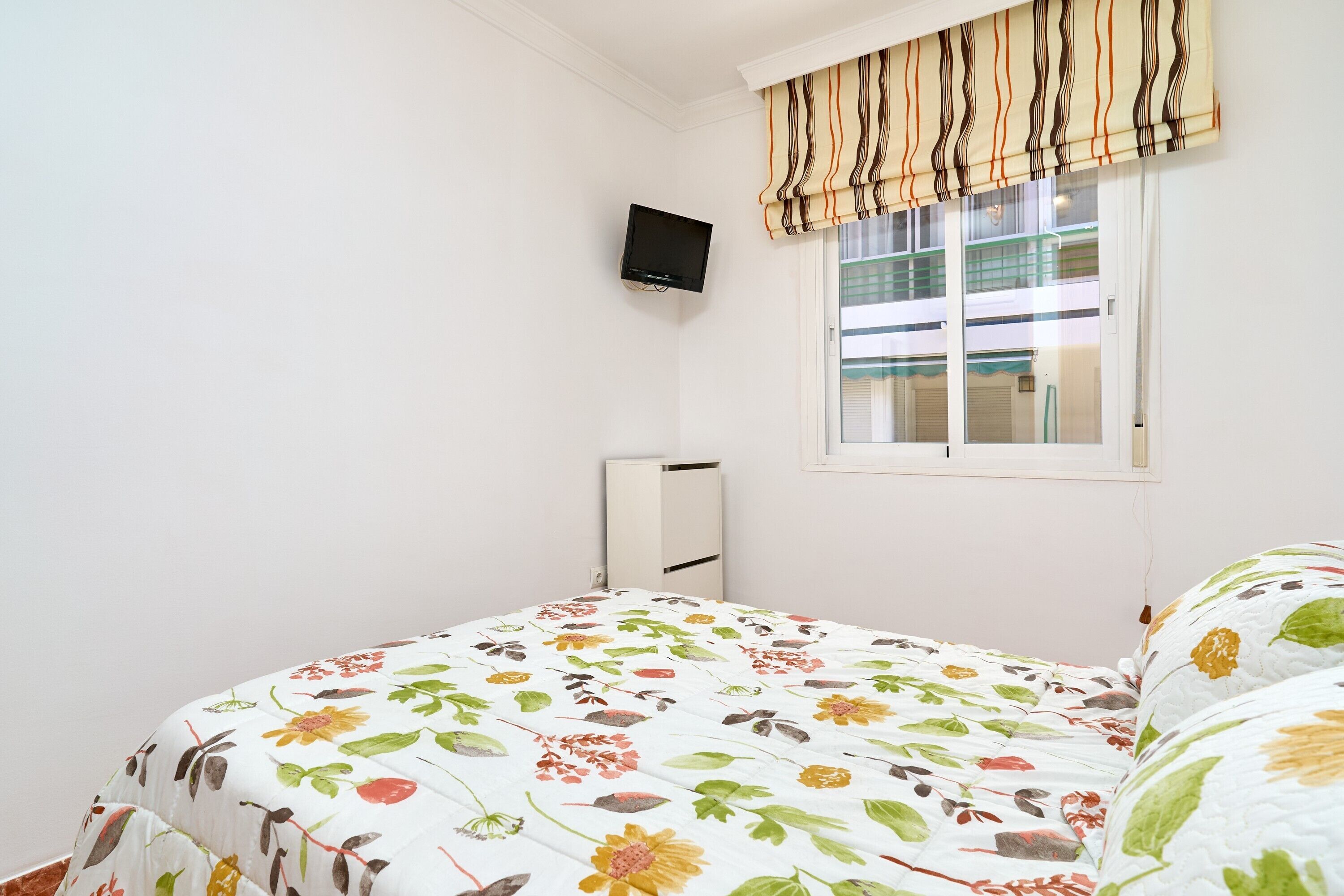Une chambre à coucher avec une couvre-lit à motifs floraux, un téléviseur fixé au mur et une fenêtre agrémentée d’une lambrequin à rayures.