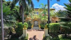 Entrada de un resort tropical con un camino pavimentado, palmeras y un edificio amarillo con una reja.