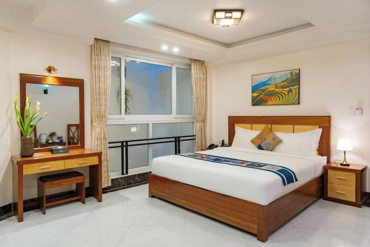 A hotel room with a bed, bedside table, nightstand, desk, chair, window, and artwork on the wall.