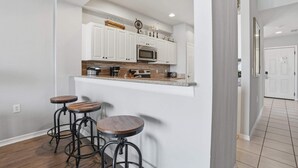 A kitchen with white cabinets, a microwave, a coffee maker, and three stools. - #914 - First Floor Golf and Lake View -Sleeps 6 (Naples)