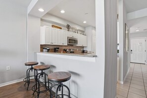A kitchen with white cabinets, a microwave, a coffee maker, and three stools. - #914 - First Floor Golf and Lake View -Sleeps 6 (Naples)