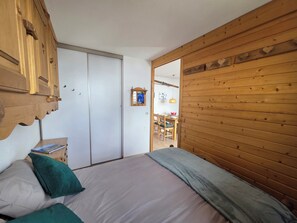 Ein Schlafzimmer mit einem Bett, Kissen, einem Holzschrank, einem Spiegel und einem Essbereich, der durch eine offene Tür sichtbar ist.