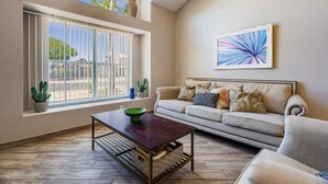 A living room with a sofa, a coffee table, a window with blinds, and a wall-mounted abstract painting. - Taylor Ranch Respite - Modern, Light, Open Concept (Albuquerque)