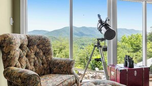 A room with a large window, a telescope, a floral armchair, and a red suitcase.