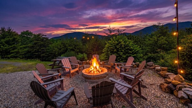 A fire pit surrounded by Adirondack chairs, set on a gravel area with a view of mountains and a twilight sky.