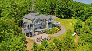 A large, multi-story house with a covered patio and a fire pit area, surrounded by dense forest.