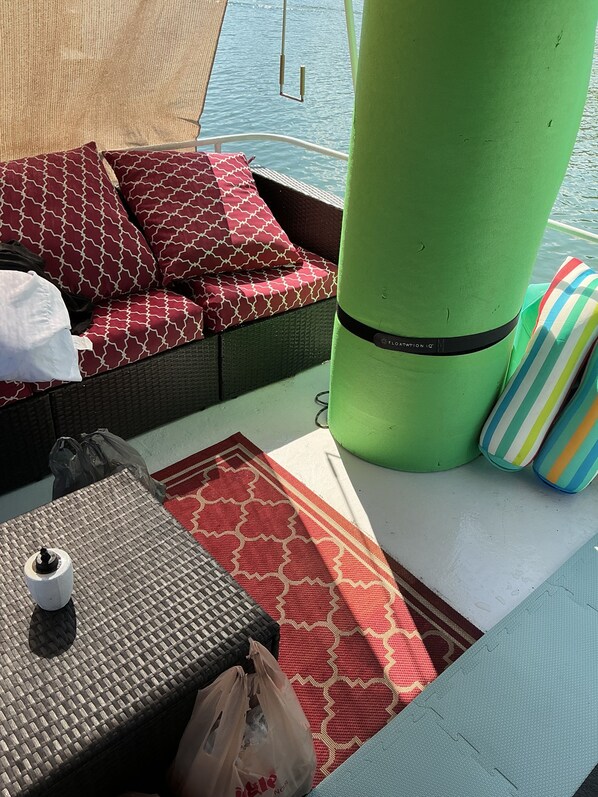 A wicker sofa with patterned cushions, a green pillar, a striped cushion, a woven table, and a plastic bag on a boat deck.