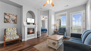 A living room with a fireplace, a television, a ceiling fan, and a view through the window. - The Golden Hour Great Views (Branson)