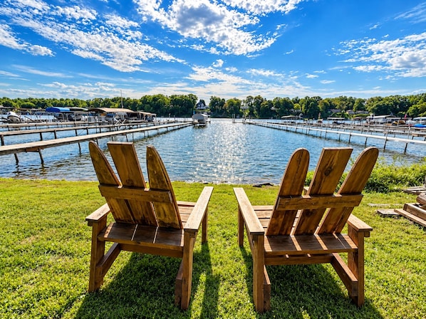 Two wooden Adirondack chairs facing a marina with boats docked, a dock extending into the water, and a clear sky above.