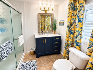 A bathroom with a glass shower enclosure, a white toilet, a dark blue vanity with gold handles, and a yellow floral shower curtain.