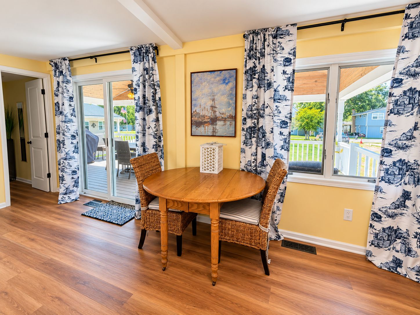 A dining area with a wooden table and wicker chairs, a painting on the wall, and a view of the outside through large windows.